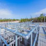 A modern outdoor water treatment facility with metal walkways, railings, and pipes under a blue sky. Trees and construction equipment are visible in the background.