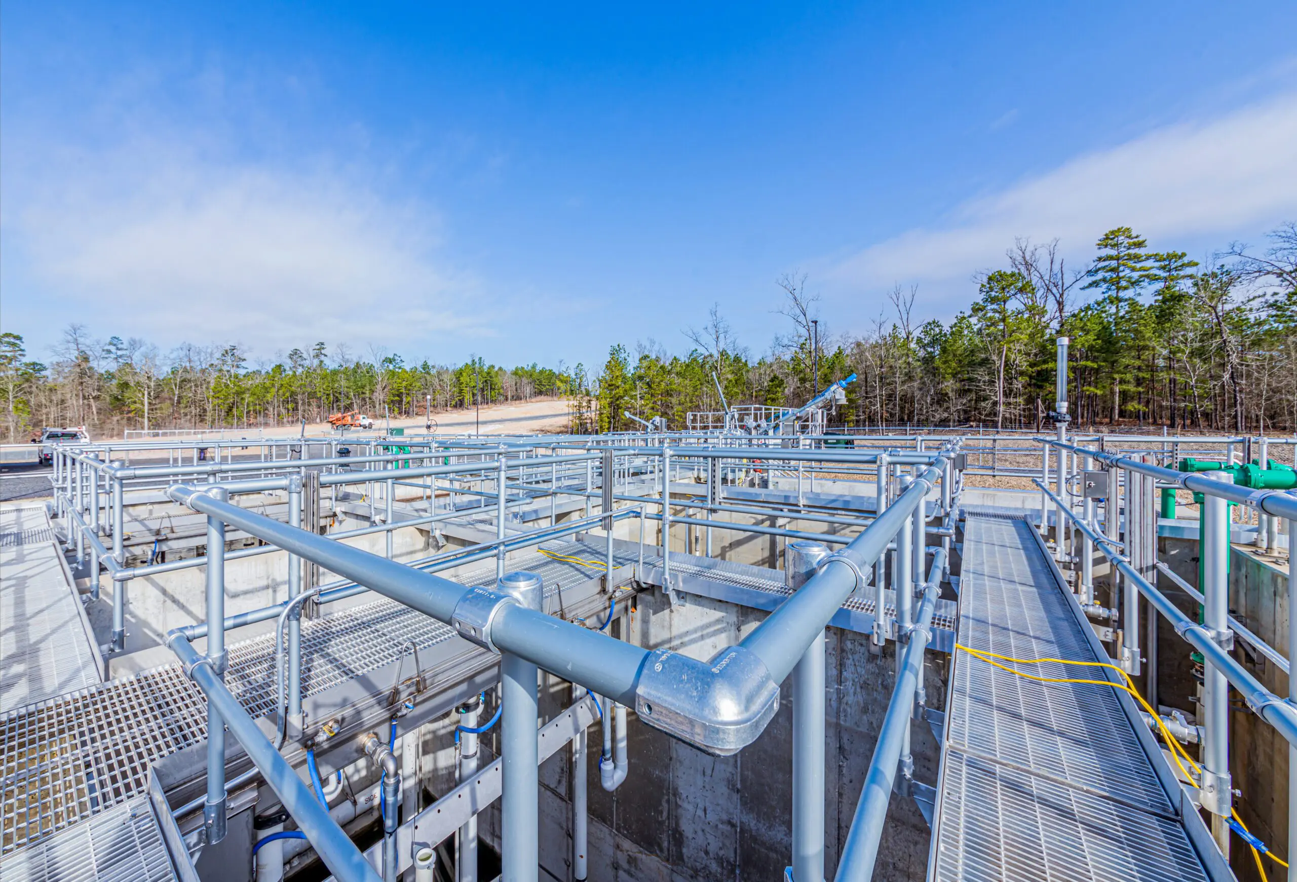 A modern outdoor water treatment facility with metal walkways, railings, and pipes under a blue sky. Trees and construction equipment are visible in the background.