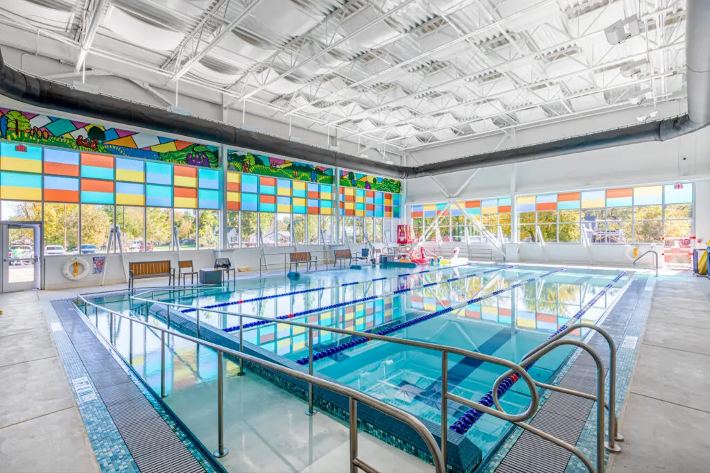 Indoor swimming pool with clear blue water, surrounded by white walls and large colorful stained-glass windows. Benches line the walls, and bright sunlight streams in from the windows.