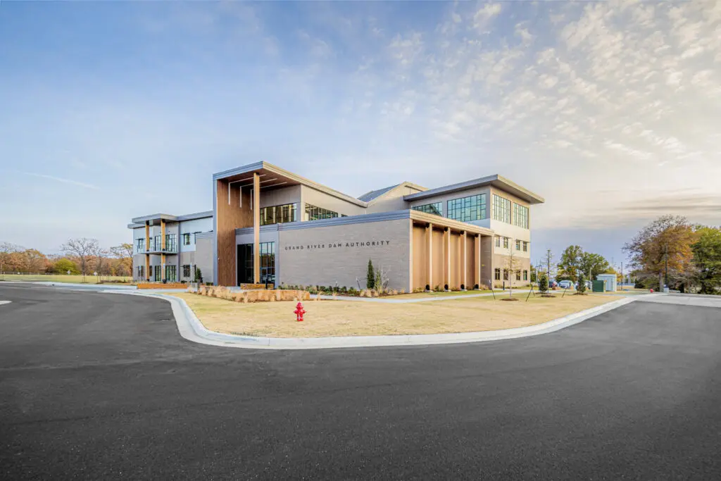 Modern two-story office building with large windows and a flat roof, surrounded by a neatly landscaped lawn, a curved driveway, and a red fire hydrant in front. The sky is partly cloudy with soft, natural light.