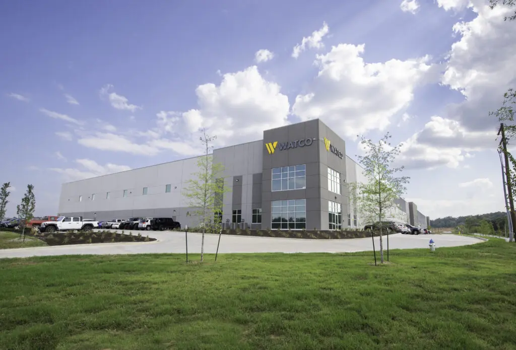 A large, modern industrial building with “WATCO” signage on the corner, surrounded by green grass, a parking lot with cars, and a partly cloudy sky.