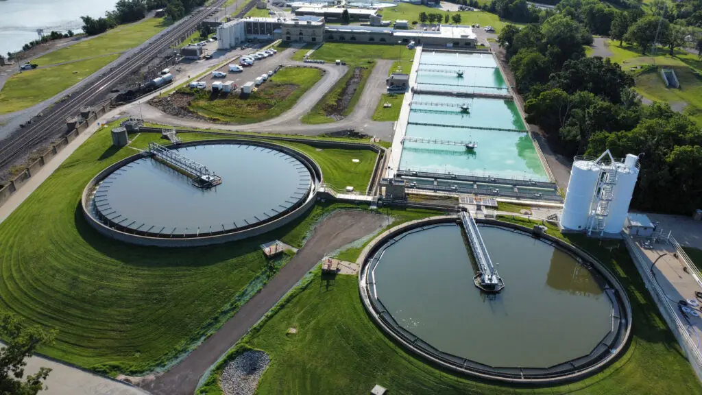 Aerial view of a water treatment plant with two large circular clarifier tanks, several rectangular settling basins, white storage silos, and adjacent green lawns surrounded by roads and industrial buildings.