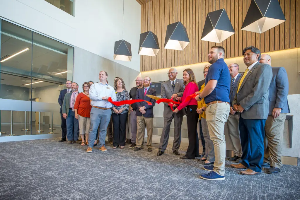 A group of people stand indoors, holding a large red ribbon for a ribbon-cutting ceremony. Some are smiling, and large geometric lights hang from the ceiling above them.