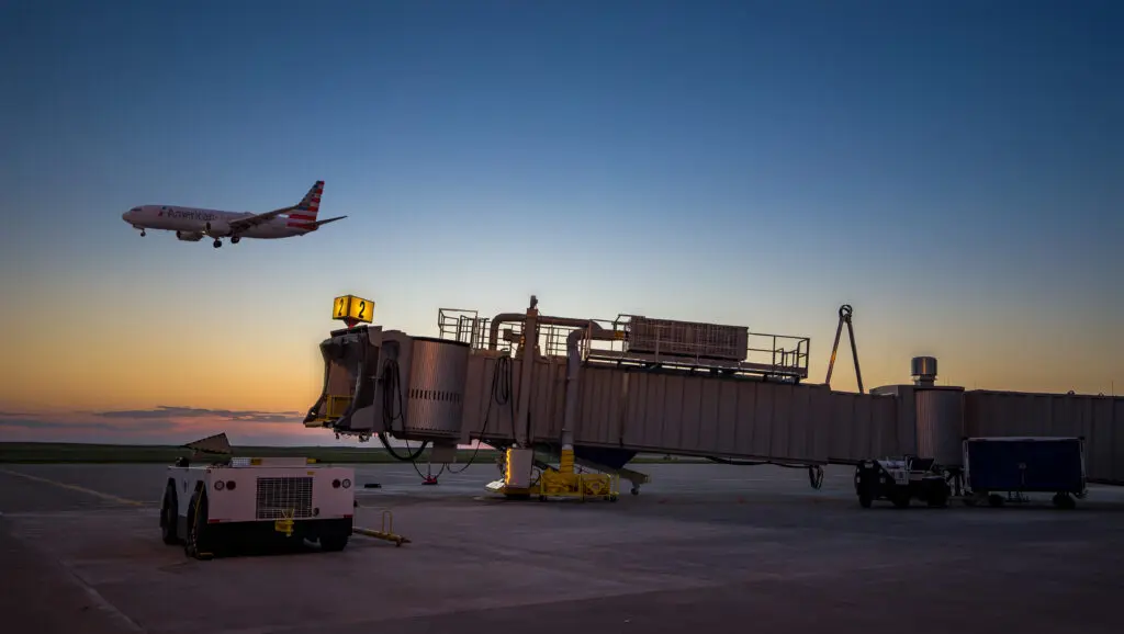 A commercial airplane prepares to land near an empty jet bridge at an airport during sunset, with ground support vehicles nearby and a clear sky in the background.