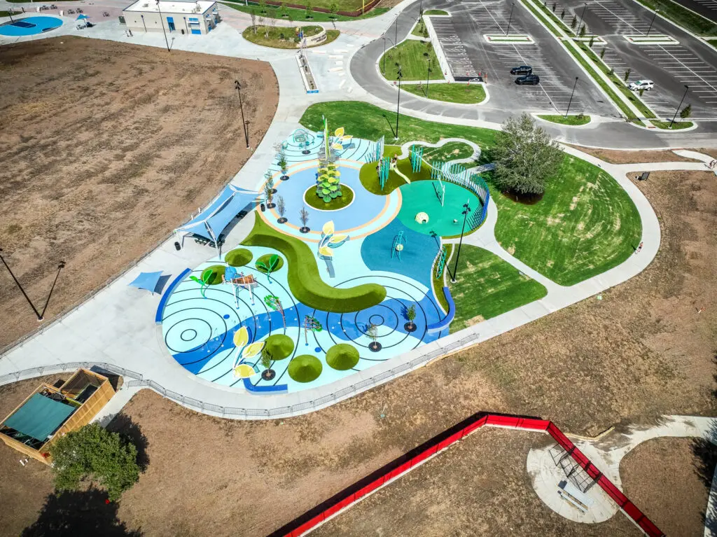 Aerial view of a colorful splash pad and playground with water features, surrounded by sidewalks, grassy areas, and parking lots, within a large park.
