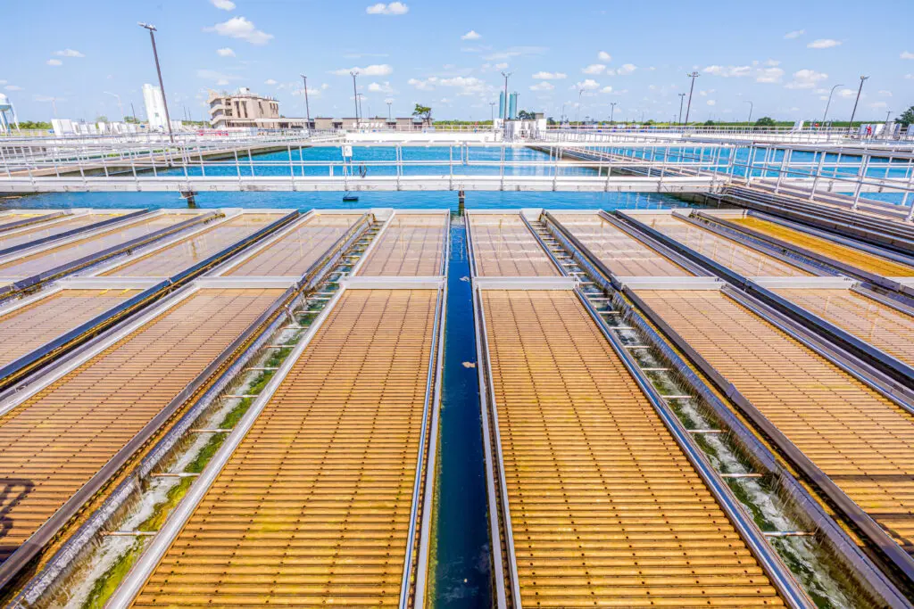 Rows of water treatment facility equipment with grates and channels, leading to large pools of clear water under a bright blue sky with scattered clouds.