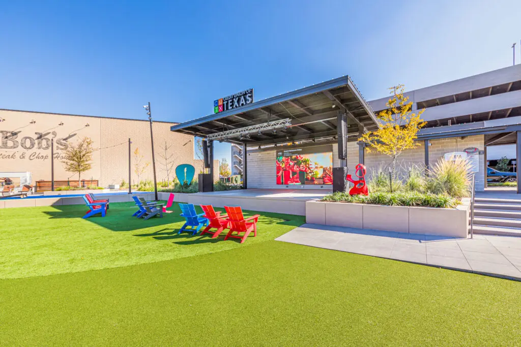An outdoor plaza with colorful Adirondack chairs on artificial grass, a stage with a large screen, and a mix of modern and rustic buildings under a clear blue sky.