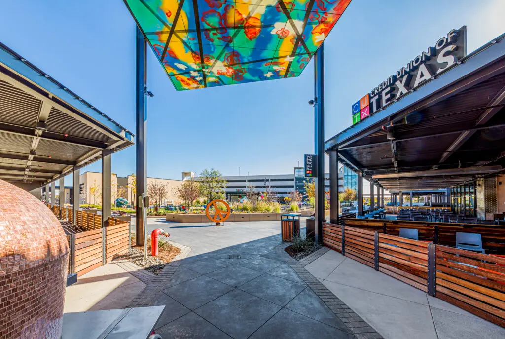 Outdoor plaza with colorful glass canopy, modern seating areas, and shaded dining spaces. A large orange sculpture is visible in the background, with a sign reading "The Sound of Texas" on a nearby building.