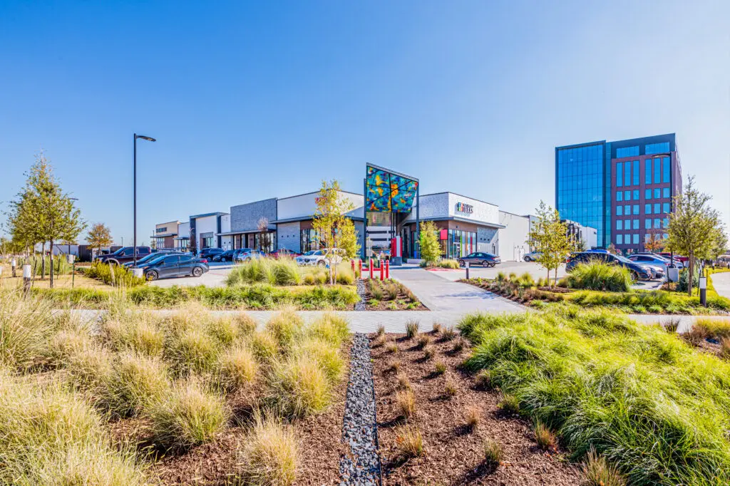 A modern shopping center with a colorful mural, surrounded by landscaped grass and plants, several parked cars, and tall office buildings in the background under a clear blue sky.