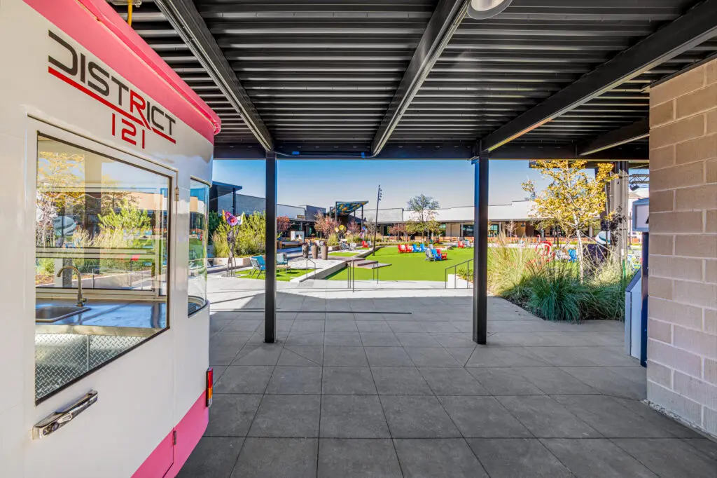 Covered outdoor walkway with a pink and white food cart labeled "District 121" on the left, leading to a courtyard with green artificial turf, seating, plants, and modern buildings in the background.