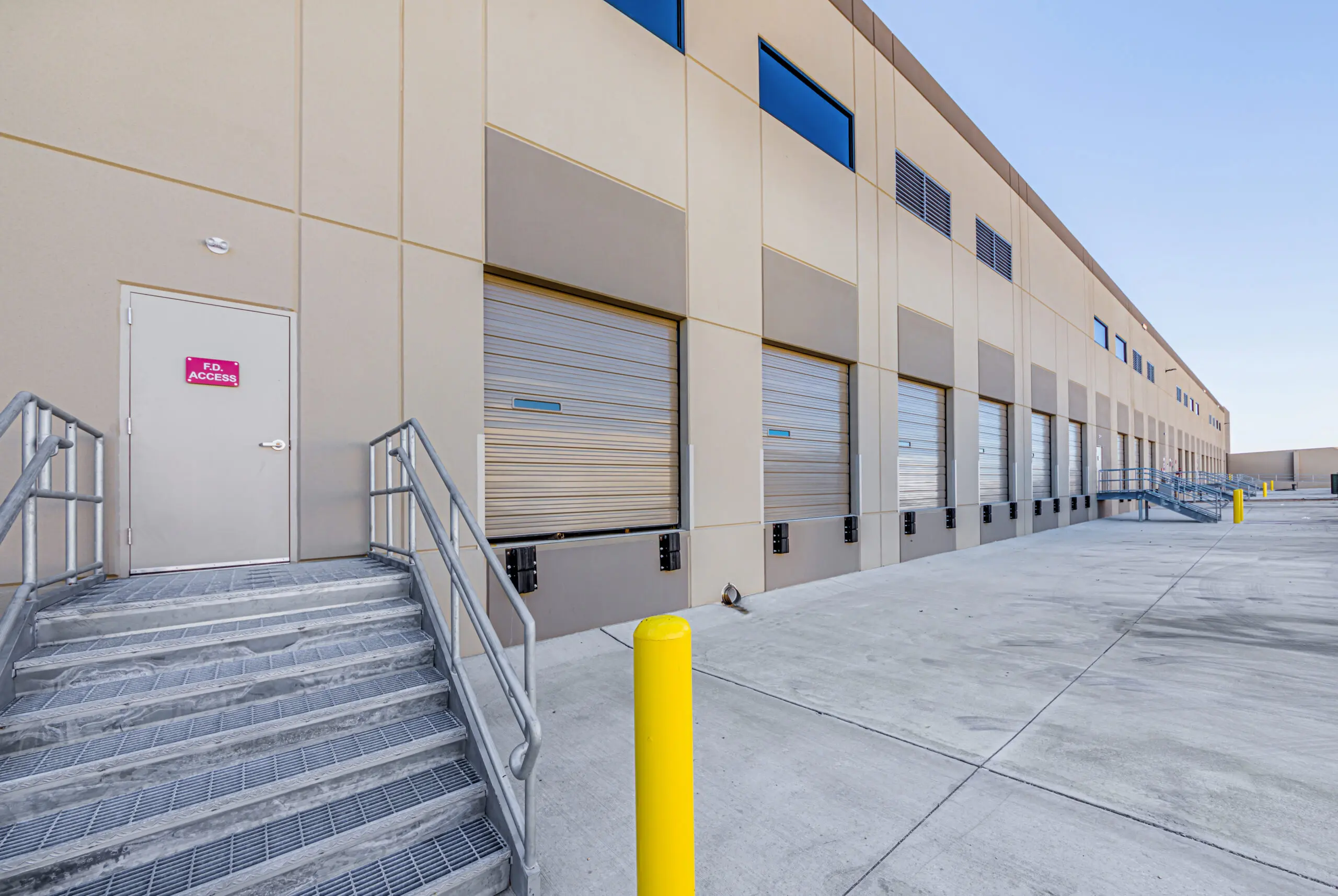 A modern industrial building with multiple closed loading dock doors, a metal staircase leading to a door marked “No Access,” and yellow safety bollards in a clean, empty concrete yard under a clear sky.