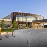 A modern building with large windows and a covered drop-off area, surrounded by landscaped greenery and a curved driveway under a clear evening sky. Bike racks and young trees are visible in the foreground.
