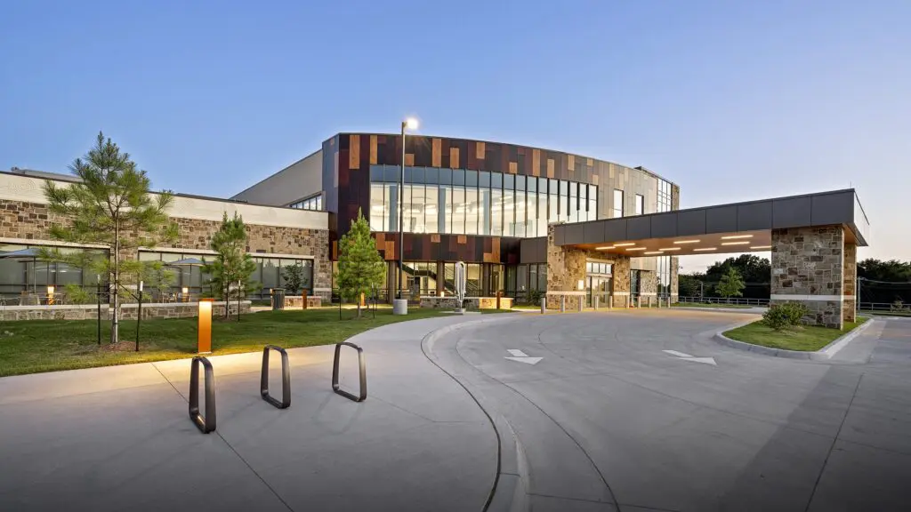 Modern building with large windows and a covered driveway, featuring stone and wood panel exteriors, surrounded by small trees and landscaped grass areas, under a clear blue sky at dusk.