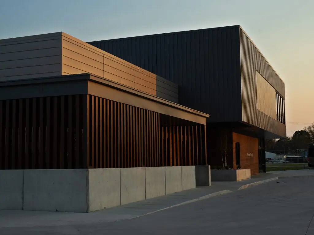 A modern building with sleek, dark metal and wood siding is pictured at sunset. The geometric design features sharp angles and large windows. Concrete barriers line the sidewalk and parking area in front of the building.