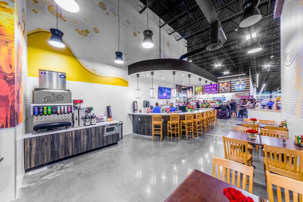 A modern, brightly lit restaurant features wooden tables and chairs, a self-serve soda fountain, and a central counter with people dining. The décor includes high ceilings, hanging lights, and wall art.