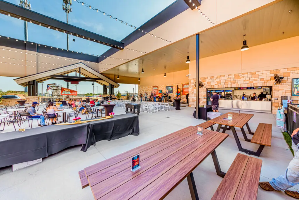 Outdoor dining area with wooden picnic tables, string lights overhead, people sitting and eating, and a counter for ordering food. The space is open-air, sheltered by a modern roof structure.