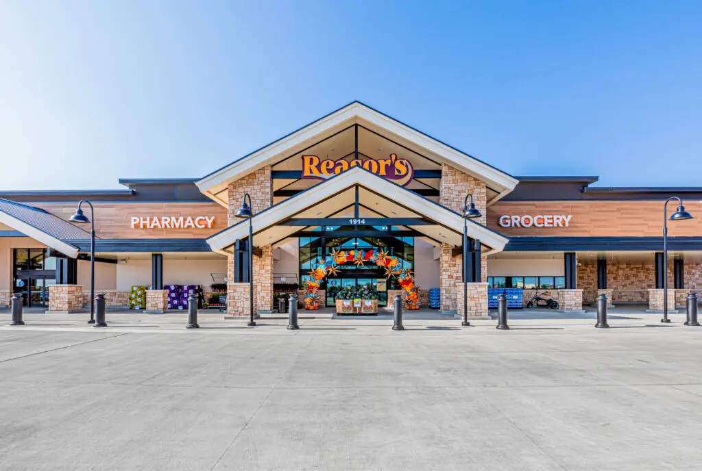 The image shows the front entrance of a grocery store named "Reasor's," with signs for "Pharmacy" and "Grocery." The entrance is decorated with a colorful display, and the sky is clear and blue.