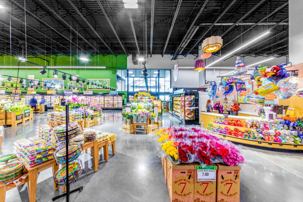 A brightly lit supermarket displays colorful flowers, candy, and produce on wooden stands. Shelves line the walls, and shoppers are visible near the entrance under high black ceilings with hanging lights.