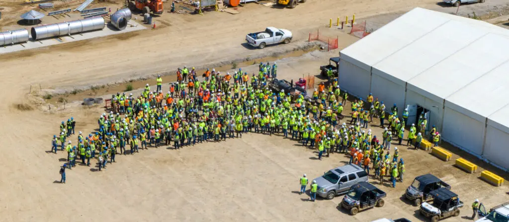 A large group of construction workers wearing safety vests and helmets gather in front of a white tent at a construction site with several vehicles and equipment visible nearby.