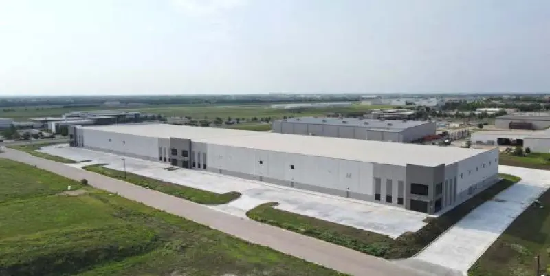 A large, white, rectangular warehouse building with multiple loading bays is surrounded by paved roads and green fields under a partly cloudy sky. Other industrial buildings are visible in the background.