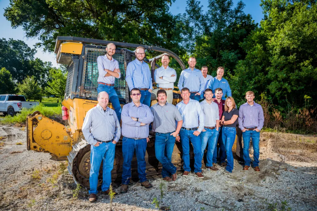 A group of fourteen people, mostly men and one woman, stand together outdoors in front of a yellow bulldozer on a sunny day. They are dressed in casual business attire, mostly jeans and button-up shirts. Trees are in the background.