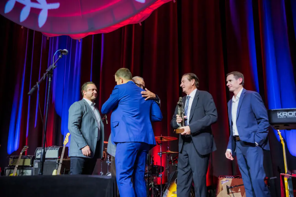 Five men in suits stand on a stage with musical instruments in the background. Two of them are hugging, while another holds a trophy. The stage is lit with blue and red lights and features a dark curtain backdrop.