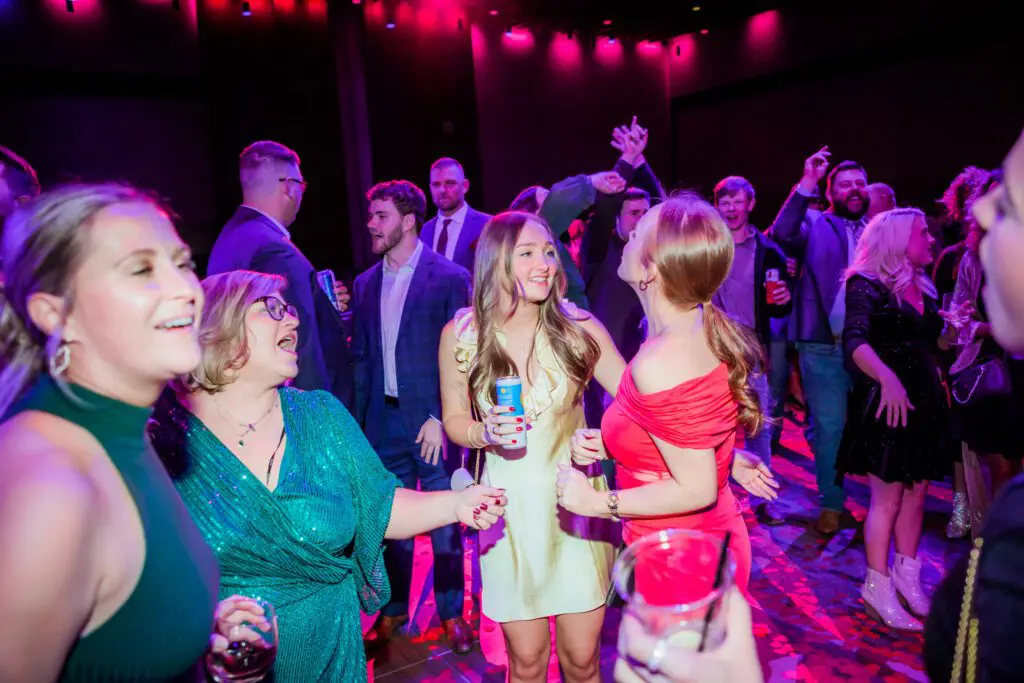 A group of people dressed in semi-formal attire dance and socialize at a lively indoor party with colorful lighting and drinks in hand.
