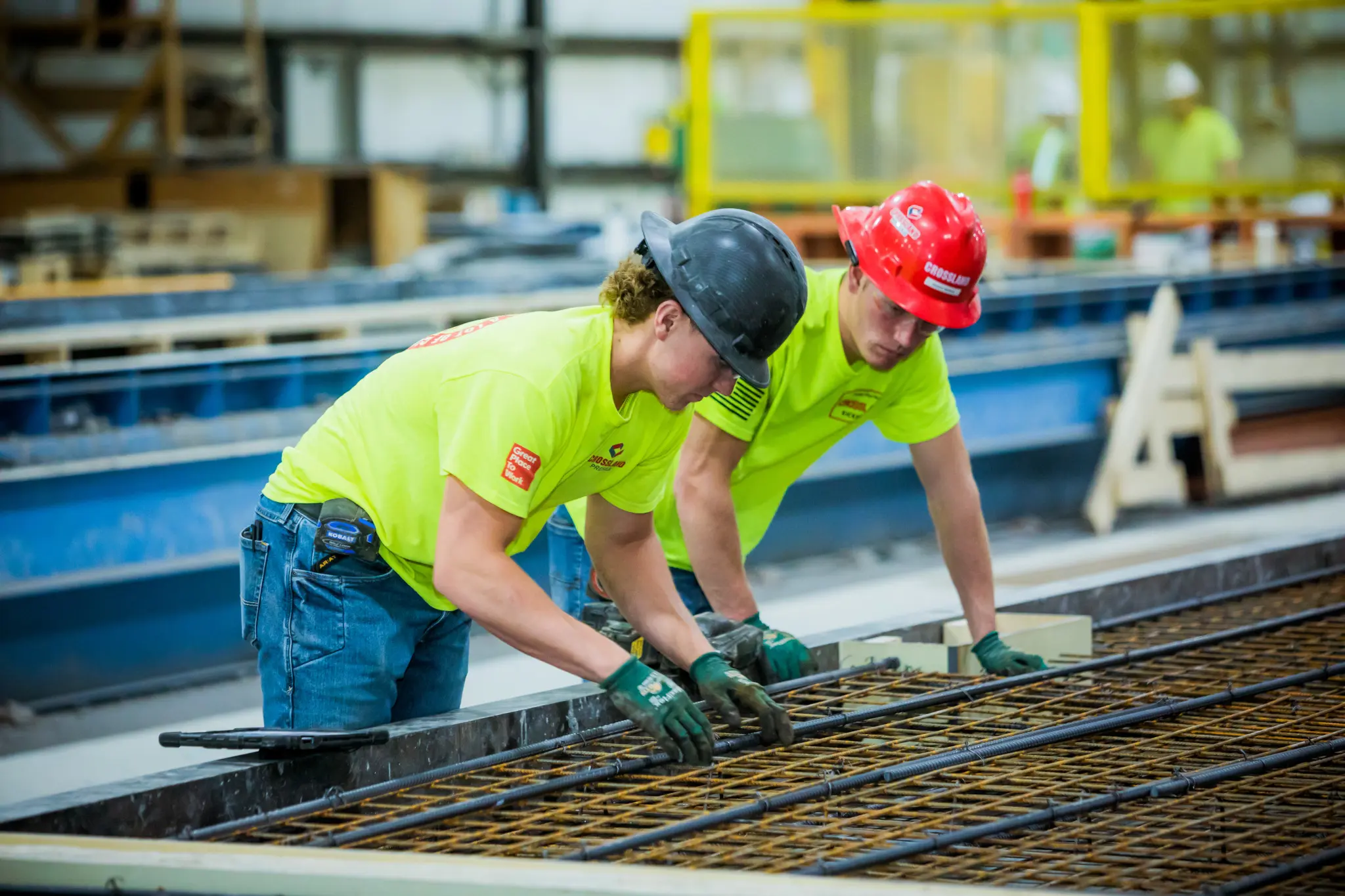 Two construction workers wearing hard hats and neon yellow shirts work together, inspecting and aligning steel rebar on a construction site indoors.