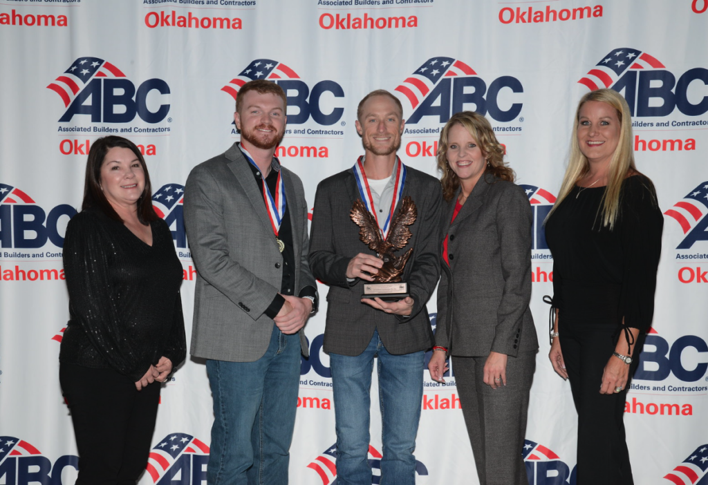 Five people stand together smiling in front of an "ABC Oklahoma" step-and-repeat banner. The person in the center holds an eagle-shaped award and wears a medal around their neck.