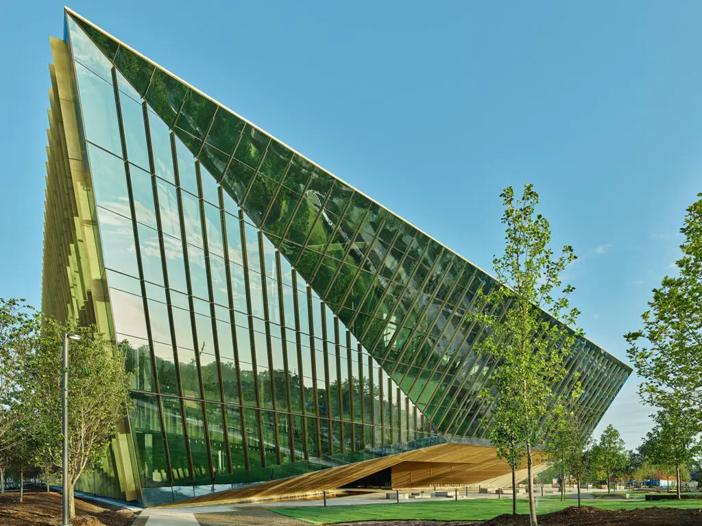 A striking modern building with tall glass walls and sharp, angular lines, set amidst a landscaped area with young trees under a clear blue sky.