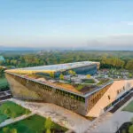 Aerial view of a modern, angular building with a green roof, surrounded by trees and landscaped grounds, under a clear sky at sunrise.