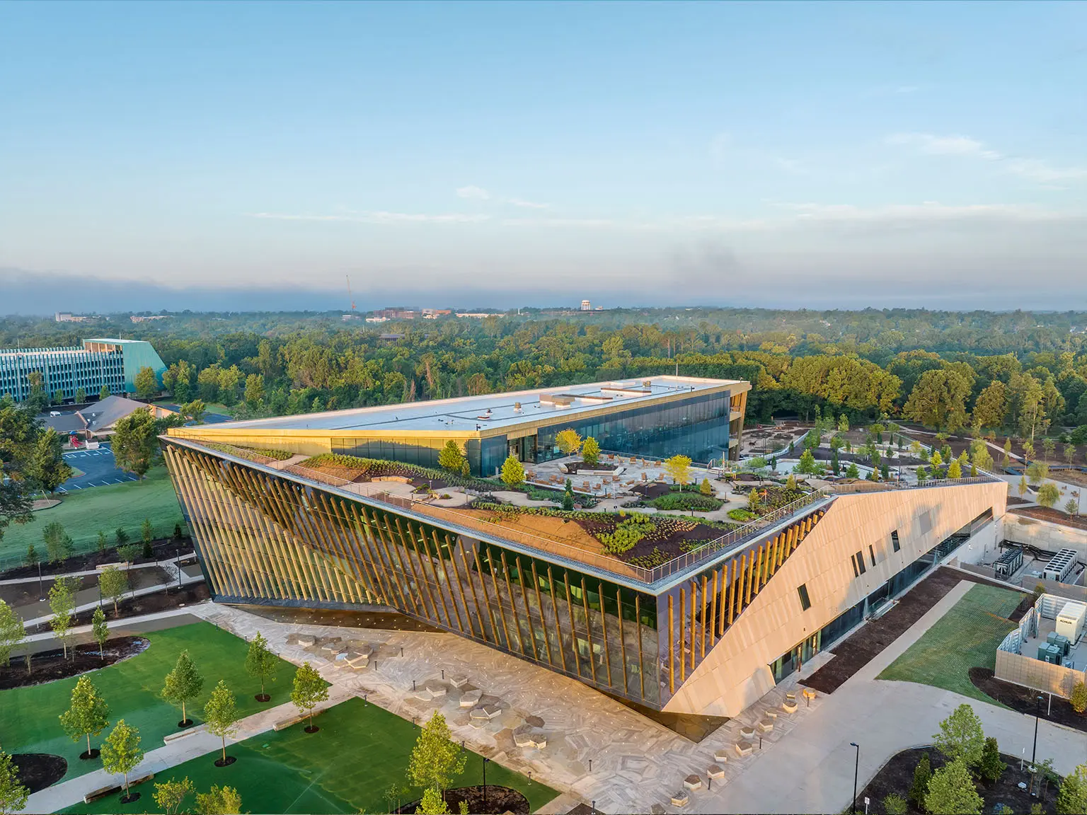 Aerial view of a modern, angular building with a green roof, surrounded by trees and landscaped grounds, under a clear sky at sunrise.
