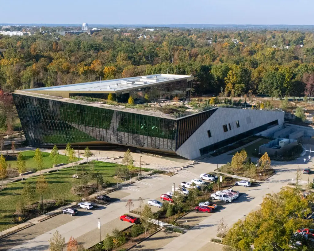 Aerial view of a modern building with angular design and reflective glass facade, surrounded by trees with fall foliage, landscaped grounds, and a parking lot with several cars.