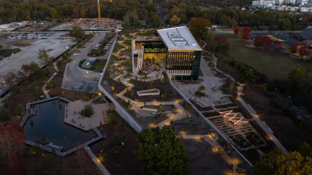 Aerial view of a modern building with large glass windows, surrounded by landscaped gardens, walkways, a pond, and illuminated pathways, set amid trees and parking areas at dusk.