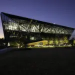 A modern, angular glass building with illuminated interiors stands surrounded by trees at dusk, with a darkening sky and a grassy area in the foreground.