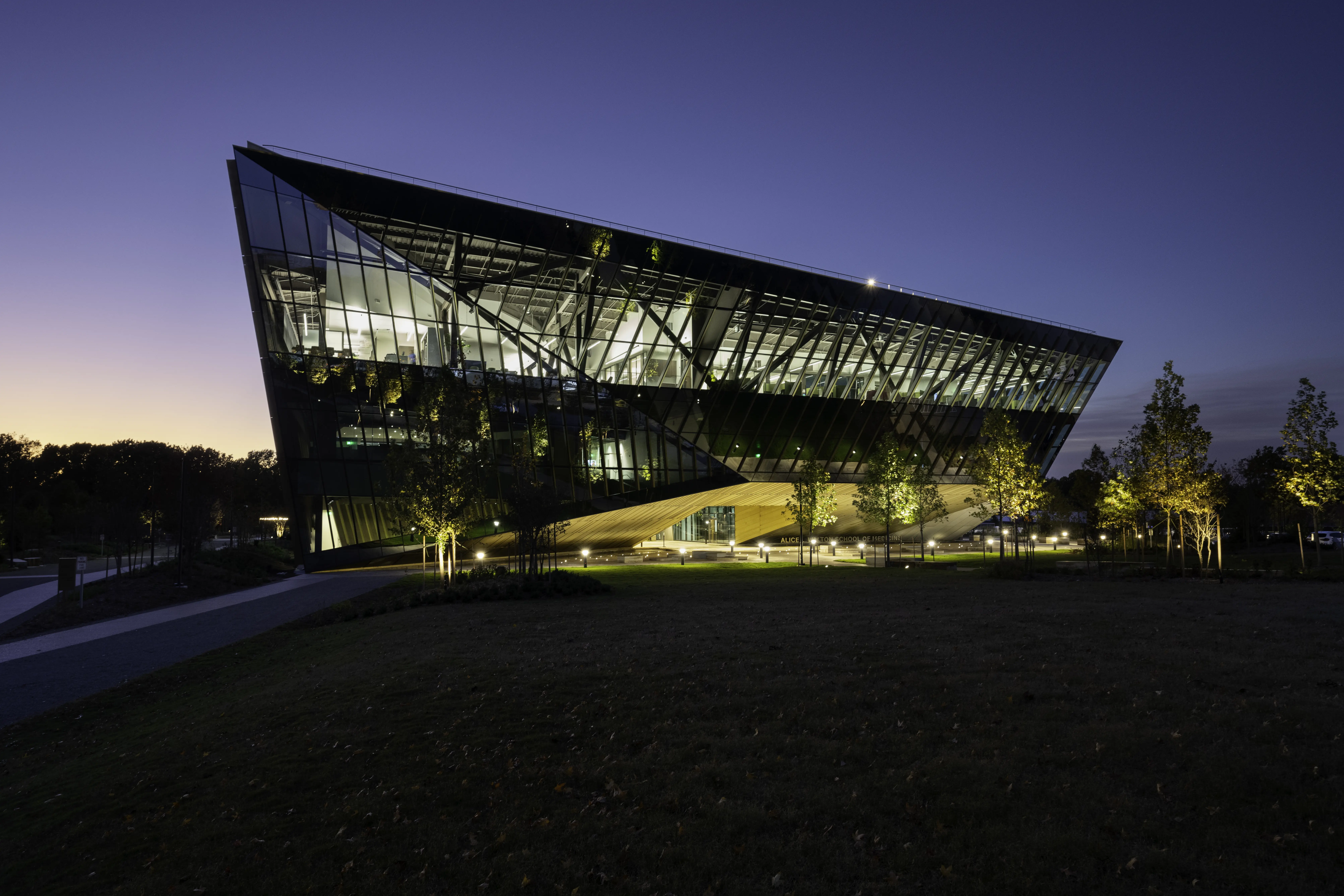 A modern, angular glass building with illuminated interiors stands surrounded by trees at dusk, with a darkening sky and a grassy area in the foreground.