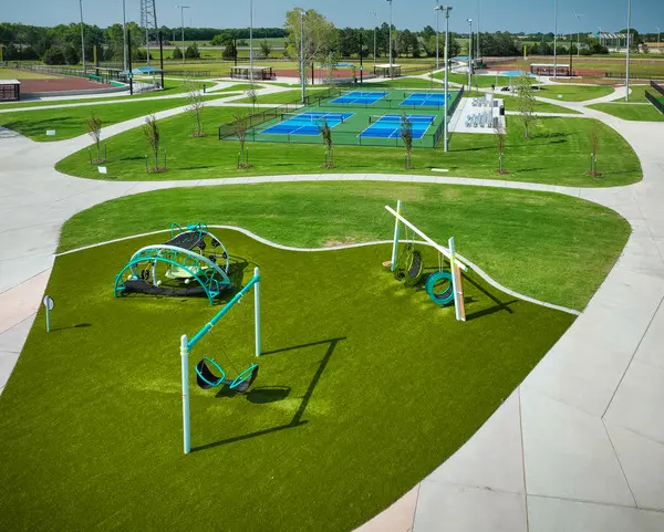 A modern park with green artificial turf, a playground with swings and climbing structures in the foreground, tennis or pickleball courts in the background, and walking paths throughout.