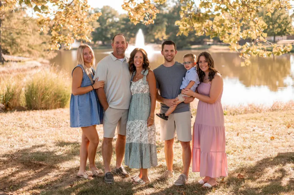 A family of six stands smiling outdoors by a lake with a fountain. They are surrounded by trees and sunlight. Three women, two men, and a young boy are dressed in casual summer clothes.