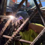 A welder wearing protective gear uses a welding torch on steel rebar, creating bright sparks. The worker is surrounded by metal rods and wears a yellow shirt with a visible logo.