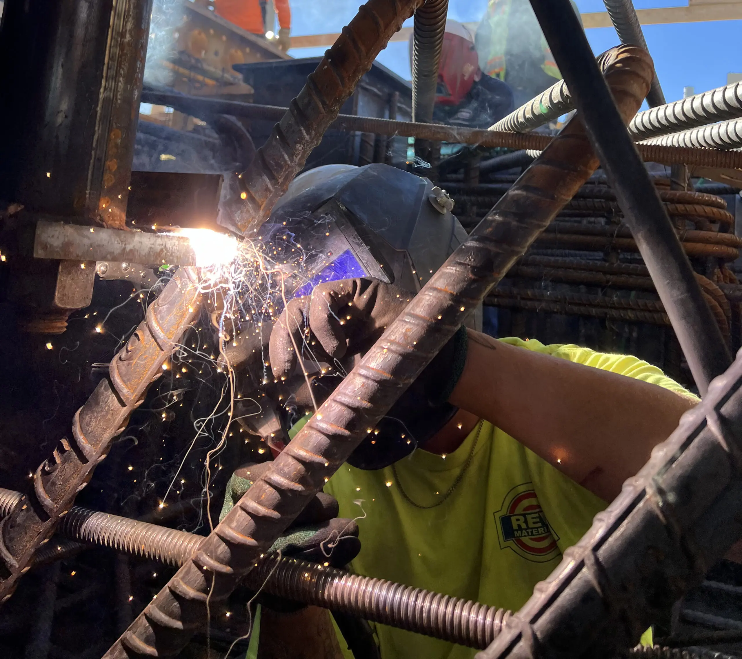 A welder wearing protective gear uses a welding torch on steel rebar, creating bright sparks. The worker is surrounded by metal rods and wears a yellow shirt with a visible logo.
