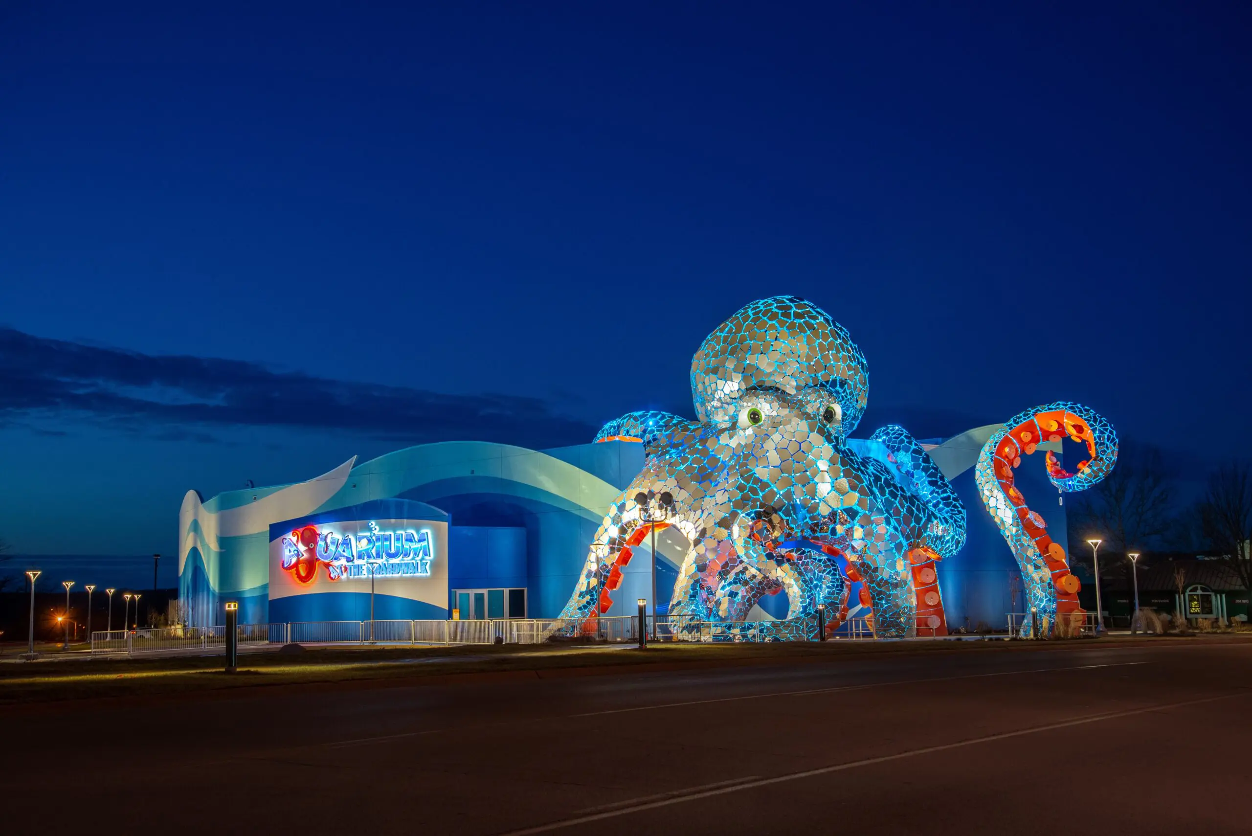 A brightly lit building at night features a giant octopus sculpture wrapped around its exterior, with blue and orange mosaic-like tiles. A glowing sign reads “Aquarium St. Petersburg.” The sky is dark blue.