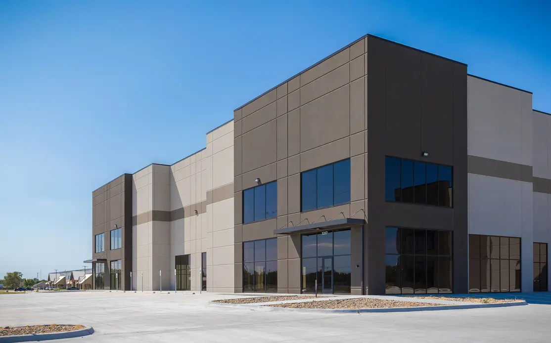 A modern, empty commercial or industrial building with large windows and a clean, paved parking lot under a clear blue sky.