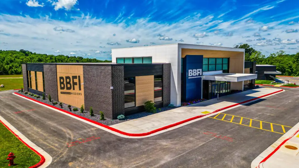 Modern building with "BBFI" signage, featuring black and wood paneling, large windows, and landscaped surroundings; set against a bright blue sky with scattered clouds and greenery in the background.