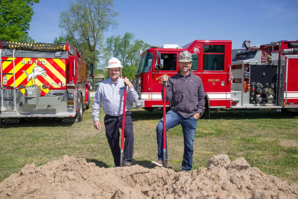 Two men wearing hard hats stand beside a dirt pile with shovels, posing in front of two red fire trucks on a sunny day with trees and blue sky in the background.