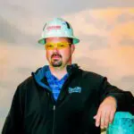 A man wearing a hard hat, yellow safety glasses, and a dark jacket stands outdoors at sunset, resting one arm on a railing. The logo "Crossland" is visible on his hat and jacket.