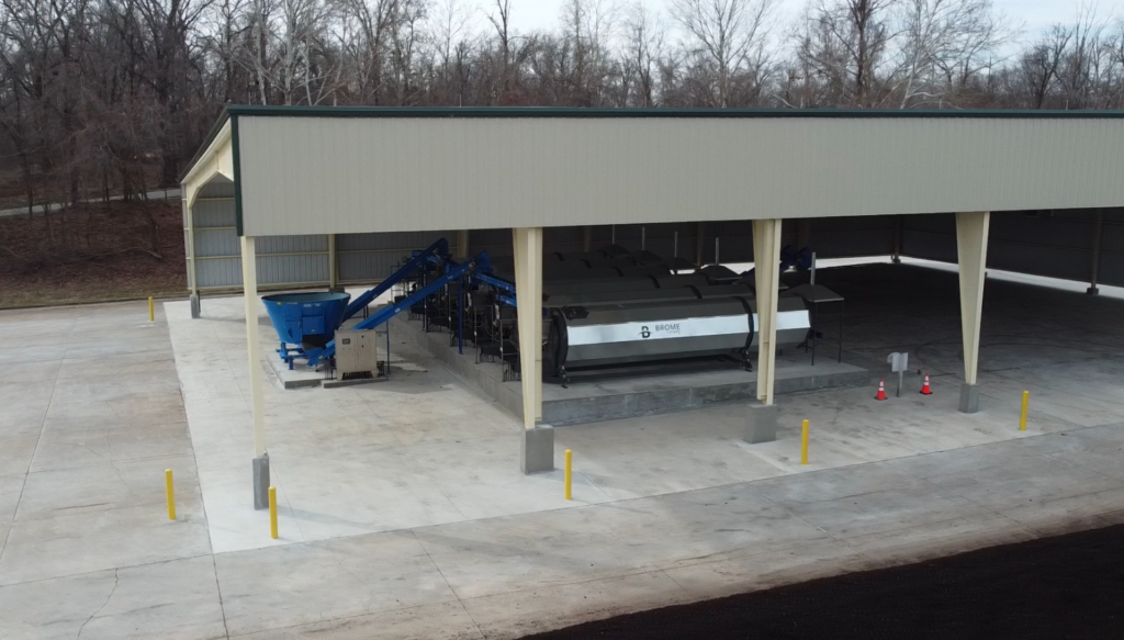 A large composting facility with industrial equipment and machinery under a metal roof, surrounded by concrete pavement. Yellow safety bollards and orange traffic cones are positioned near the machines. Leafless trees are in the background.
