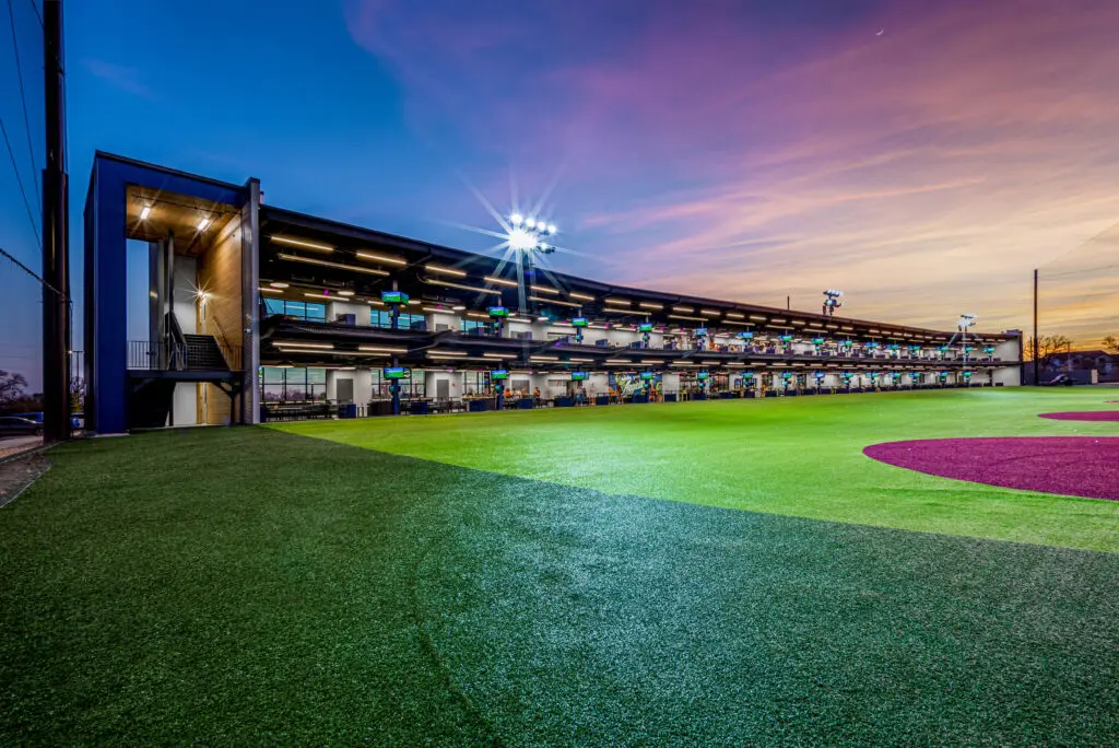 A modern multi-level driving range with bright lights overlooks a well-maintained artificial turf field at sunset, with a colorful sky in the background.