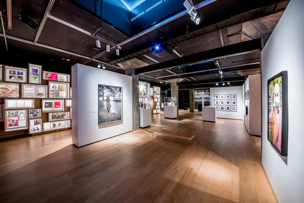 A modern art gallery with wooden floors, white walls displaying framed photographs and art, and spotlights on the ceiling. The space has benches and a clean, open layout with art on every wall.