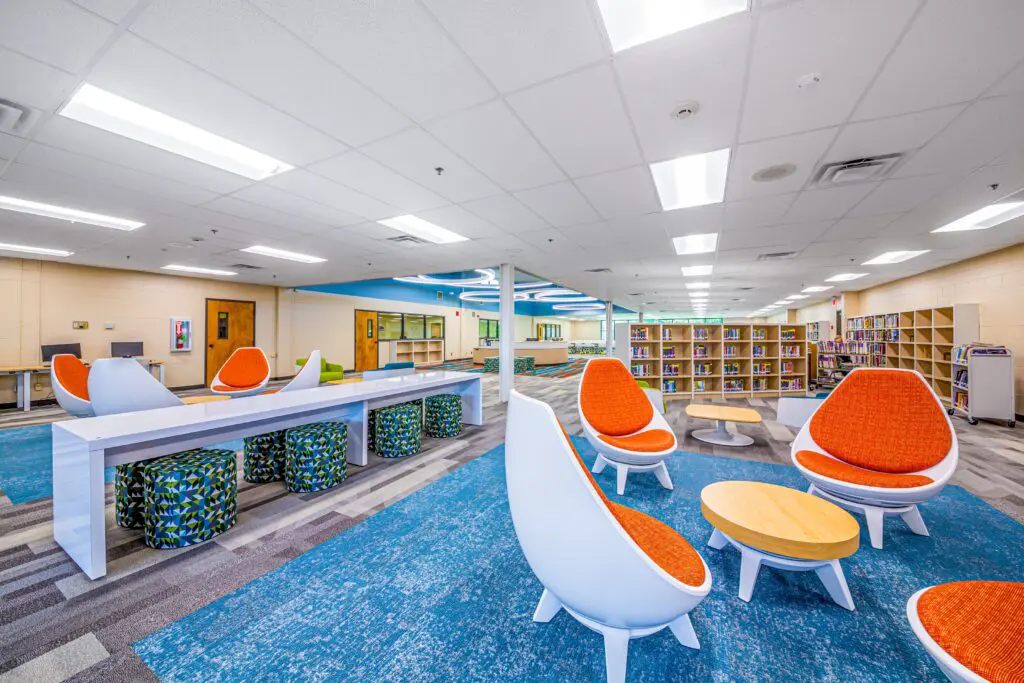 A modern, brightly lit library features orange and white chairs, small wooden tables, long desks with patterned stools, and bookshelves filled with books along the walls. The carpet is blue and gray.