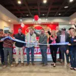 A group of people smile and cheer as they cut a blue ribbon at an indoor ribbon-cutting ceremony, with balloons and a red wall in the background.